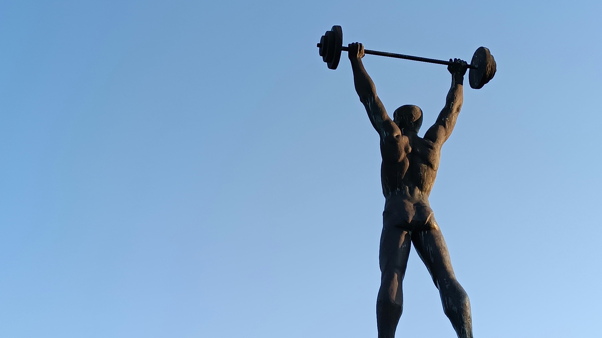 A bronze statue of a muscular figure hoisting a barbell overhead, seen from behind against a clear blue sky, legs planted and straining under the load